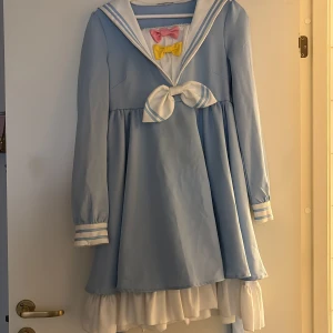 Sailorklänning från Kawaii.se, blå/vit S - Supersöt ljusblå sailorklänning med vit krage och volangkant, dekorerad med avtagbara rosetter i rosa, gul och vit. Långärmad modell med knytrosett framtill och klassiska ränder på ärmslut och krage. Perfekt för dig som älskar kawaii-stil! 