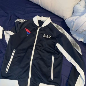 EA7 svart och vit vindjacka - Sportig svart och vit vindjacka från EA7 med vita ränder på axlarna, dragkedjor på fickor och framtill. Jackan har en snygg logga på bröstet och färgdetalj i rött och blått. Perfekt för en aktiv och stilren look.