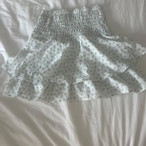 Vit blommig volangkjol med smock - Supersöt kort kjol i vitt med små blommor och volanger. Kjolen har bred smock i midjan som ger en bekväm och snygg passform. Perfekt för varma dagar och lätt att matcha med en enkel topp.