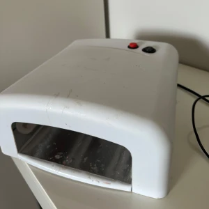 Vit UV-lampa för naglar - UV-lampa i vit plast med svart och röd knapp, perfekt för att härda gelnaglar hemma. Lampan har en klassisk rektangulär form och plats för hela handen. Kopplas in med sladd. Gör din nagelrutin smidigare!