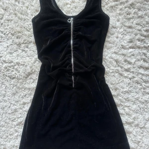 Svart sammetsklänning Red Label - Supersnygg svart kort sammetsklänning från R&D Red Label. Klänningen har en dragkedja framtill med ett hjärtformat hänge och rynkade detaljer längs dragkedjan. Ärmlös och figurnära modell.