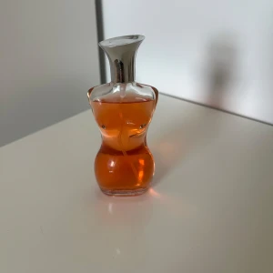 Parfym - Ikonisk parfymflaska formad som en kvinnokropp. Flaskan är i glas med silvrig metallkork och innehåller en orangefärgad doft. Perfekt för dig som gillar unika och snygga flaskor på hyllan.
