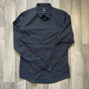 H&M svart slim fit skjorta - Stilren svart skjorta från H&M i slim fit-modell. Skjortan har storleken M. Den är i jättebra skick. Längden 81 cm, axeln 45 cm