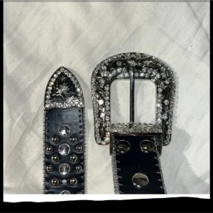 Svart glittrigt skärp med strass - Säljer ett svart bälte med massor av glittrande strass och stora silverfärgade nitar. Spännet är dekorerat med bling och har snygga detaljer. Perfekt accessoar för att lyfta vilken outfit som helst med extra edge och glamour.