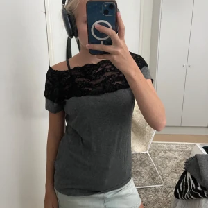 Grå topp med svart spetsdetalj - Snygg och trendiga grå topp från H&M med bred svart spets upptill som går över axlarna. Offshoulder-modell med korta ärmar och mjukt, stretchigt material. 