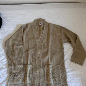 Beige stickad kofta från Doğa - Beige stickad kofta från Doğa med fina virkade detaljer längs kanterna, knappar framtill och två fickor. Koftan har diskret mönsterstickning och långärmad passform. Perfekt för lager-på-lager och chill vibes.