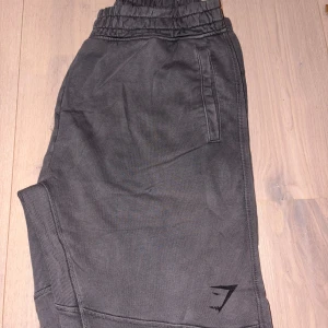 Grey Power Joggers från Gymshark - Säljer ett par grå Power joggers från Gymshark med elastisk midja och muddar vid benslut. Byxorna har en relaxed fit, sidofickor och diskret logga på benet. Perfekta för chill eller gymmet, riktigt sköna och stilrena.