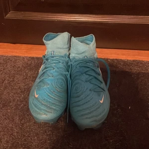 Nike Phantom Luna blå fotbollsskor elite - Nike Phantom Luna fotbollsskor i en snygg blå färg med vita detaljer och Nike-logga på sidan. Skorna har hög strumpliknande krage, snörning och mönstrad ovandel för extra grepp. Perfekta för dig som vill sticka ut på planen. Påse ingår 