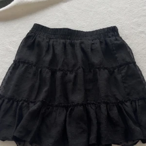 Svart prickig volangkjol från Sisters Point - Supersöt svart kortkjol från Sisters Point med volanger och prickigt mönster. Kjolen har resår i midjan för skön passform och är gjord i ett lätt, flowy material som ger en cool vibe. Perfekt att styla med sneakers eller boots för en trendig look.