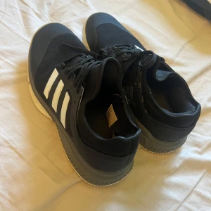 Svarta Adidas sneakers med vita ränder - Svarta Adidas sneakers med klassiska vita ränder på sidorna och grå detaljer vid sulan. Ovandelen är i mesh och syntet, vilket ger en sportig vibe. Skorna har snörning och en platt sula som passar perfekt till en aktiv sport, har använts till innebandy. 