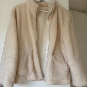 Beige teddyjacka med dragkedja - Mysig beige teddyjacka med dragkedja framtill och två stora fickor. Jackan har en hög krage och är tillverkad i fluffigt teddy-material som håller dig varm. Perfekt för kyliga dagar och enkel att matcha med olika outfits.