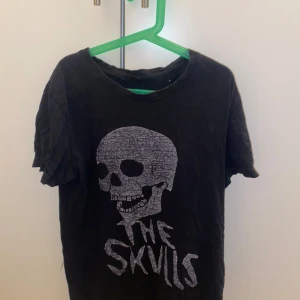 Svart t-shirt med dödskalletryck - Cool svart t-shirt med stort vitt dödskalletryck och texten 'THE SKVLLS' på framsidan. Klassisk rund halsringning och korta ärmar. Perfekt för dig som gillar rockig stil och statement-plagg. Den har ett litet hål på backsidan