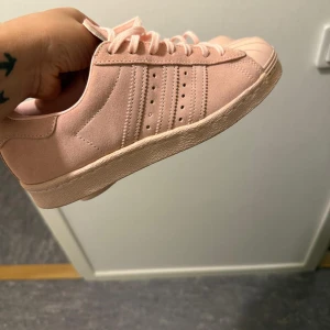 Adidas Superstar rosa mocka sneakers - Snygga Adidas Superstar sneakers i ljusrosa mocka med klassiska tre ränder på sidan och snörning. Skorna har den ikoniska gummitåhättan och platt sula. Perfekta för dig som vill ha en stilren och trendig look med en touch av färg.