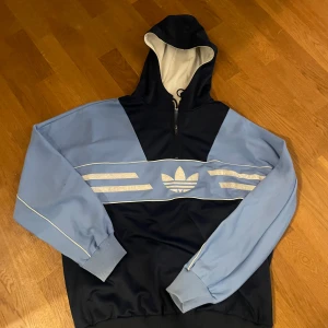 Adidas blå hoodie med half zip - Snygg hoodie från Adidas i ljusblått och mörkblått med klassiska trefoil-loggan framtill. Tröjan har huva, half zip och långa ärmar med ribbade muddar. Perfekt för dig som gillar sportig och avslappnad stil.