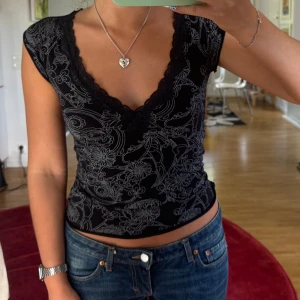 Svart blommig topp med spetskant - Supersnygg svart topp med blommönster och djup v-ringning. Spetskant längs urringningen ger en extra detalj. Modellen är tight och ärmlös, perfekt till jeans eller kjol. Stretchigt material som sitter snyggt på kroppen.