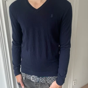Mörkblå merinoullströja Polo Ralph Lauren - Snygg mörkblå tröja från Polo Ralph Lauren i tvättbar merinoull. Slim fit-modell med v-ringning och diskret logga på bröstet. Perfekt för dig som gillar stilrena och klassiska plagg med en modern passform.