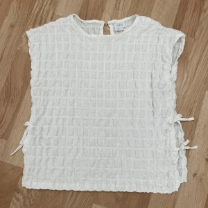  blus från Zara - Superfin vit blus från Zara med rutigt strukturerat tyg och knytband i sidorna. Modellen är ärmlös med rund halsringning och liten öppning bak i nacken. Den är i storlek 140🥰