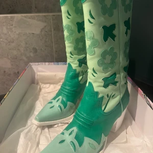 Mintgröna cowboy boots Jeffrey Campbell - Unika cowboy boots i mintgröna och gröna toner från Jeffrey Campbell. Skorna har blommiga detaljer, spetsig tå och vit klack. Tillverkade i skinn med coola utskurna mönster och retrovibbar. Perfekta för dig som vill sticka ut med färg och stil.