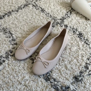 Beige ballerinaskor med rosett - Snygga beige ballerinaskor från H&M med rund tå och liten rosett framtill. Skorna har en diskret textur i materialet och platt sula, perfekta för en stilren och enkel look. Passar till både jeans och klänning. Aldrig använda. 