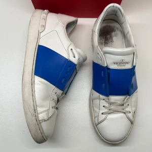 Valentino open - Snygga Valentino Garavani Open sneakers i vitt skinn med en bred blå rem över mitten. Klassisk rund tå, platt sula och snörning framtill. Märkeslogga på plösen och yttersulan. Perfekta för dig som gillar statement-sneakers med lyxig känsla.