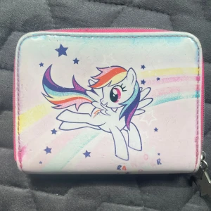 My Little Pony plånbok med regnbåge - Söt plånbok med My Little Pony-motiv och regnbågsfärger. Plånboken är ljusrosa med stjärnor och har dragkedja med stjärnformad detalj. Perfekt för att förvara kort och mynt, och superfin för dig som gillar färgglada accessoarer.