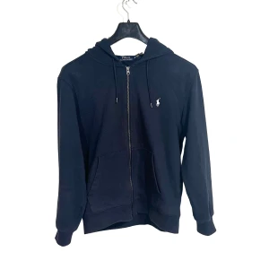 Svart hoodie från Polo Ralph Lauren - Klassisk svart hoodie från Polo Ralph Lauren med dragkedja, huva och snörning. Tröjan har en vit broderad logga på bröstet och två fickor framtill. Tillverkad i mjuk bomull för en skön och avslappnad stil.