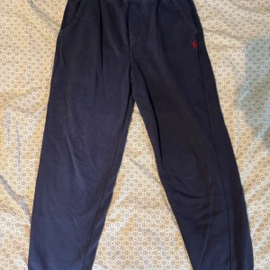 Mörkblå mjukisbyxor Polo Ralph Lauren - Mörkblå mjukisbyxor från Polo Ralph Lauren med klassisk röd logga broderad på ena fickan. Byxorna har resår i midjan och är tillverkade i mjukt bomullsmaterial. Perfekta för chill dagar eller när du vill vara bekväm men ändå se snygg ut.