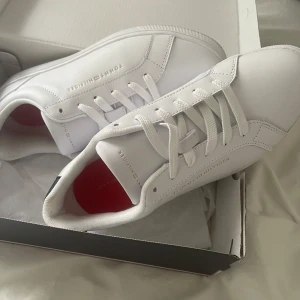 Vita sneakers från Tommy Hilfiger - Stilrena vita sneakers från Tommy Hilfiger med diskret logga i guld på sidan och tungan. Skorna har klassisk snörning, rund tå och en svart detalj på hälen. Ovandelen är i slätt syntetmaterial och innersulan är röd för en snygg kontrast. De är helt nya från Zalando, jag har bara använt dem en gång 
