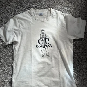 Vit C.P. Company t-shirt med print - Vit t-shirt från C.P. Company i mjuk bomull. Framsidan har ett stort svart tryck med en tecknad figur och loggan. Klassisk rund hals och korta ärmar. Perfekt för dig som gillar streetwear och snygga prints. Passar även M