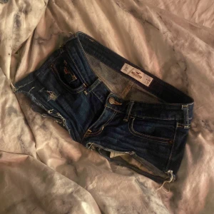 Mörkblå jeansshorts från Hollister - Snygga mörkblå jeansshorts från Hollister med låg midja och slitna detaljer. Klassisk femficksmodell med råa kanter och gul kontrastsöm. Perfekta för varma dagar och chill stil.