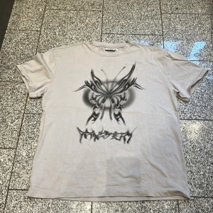 Beige oversized t-shirt från Weekday - Snygg beige t-shirt från Weekday i oversized passform. Framsidan har ett stort grafiskt tryck med en tribal-inspirerad fjäril och texten 'Misery' under. T-shirten är kortärmad och gjord i mjuk bomull, perfekt för dig som gillar streetwear och coola prints.