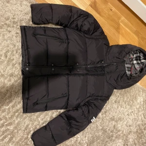 Svart pufferjacka från Burberry - Säljer en svart pufferjacka från Burberry med huva och dragkedja framtill. Jackan har ett rutigt innerfoder i grått, vitt och rött samt Burberry-logga på ärmen. Perfekt för kalla dagar och riktigt snygg streetstil.