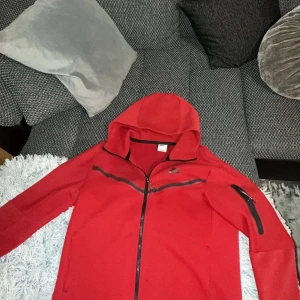 Röd Nike hoodie med dragkedja - Säljer en röd hoodie från Nike med hel dragkedja och huva. Tröjan har svarta detaljer, inklusive en dragkedja på ärmen och en diagonal dragkedja framtill. Perfekt för dig som gillar sportig stil och vill sticka ut med färg.