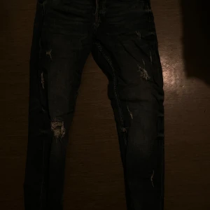 Mörka slitna jeans från Zara - Säljer ett par mörkblå jeans från Zara med snygga slitningar och trasiga detaljer på benen. Jeansen har klassisk femficksdesign och raka ben. Perfekta för dig som gillar en lite ruffare streetstil.