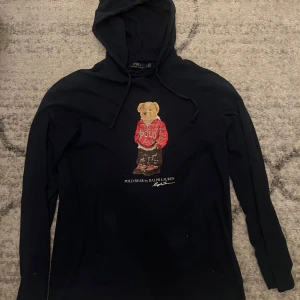 Svart hoodie Polo Bear Ralph Lauren - Marinblå  hoodie från Ralph Lauren med Polo Bear-tryck på bröstet. Tröjan har huva med snörning och lång ärm. Materialet är mjuk bomull och designen är klassisk med en cool björn i röd hoodie framtill. Perfekt för dig som gillar streetwear och ikoniska prints.