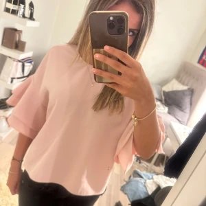 Ljusrosa blus med volangärmar - Supersöt ljusrosa blus från Zara med breda volangärmar och rund halsringning. Blusen har en loose fit och stängs med en liten knapp i nacken. Perfekt för dig som gillar en feminin och trendig look.