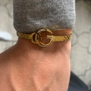 Gucci guldigt armband med GG-logo - Snyggt armband från Gucci i guldigt metall med klassisk GG-logo framtill. Armbandet har ett diskret mönster längs bandet och är prytt med små detaljer runt loggan. Perfekt för dig som vill ha en lyxig och trendig accessoar.