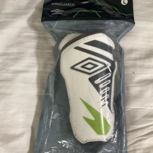 Umbro benskydd vit/svart/grön L - Säljer ett par Umbro benskydd i storlek L. De är vita med svarta och limegröna detaljer och har en sportig design med Umbro-loggan på framsidan. Perfekta för dig som spelar fotboll och vill ha bra skydd på planen.