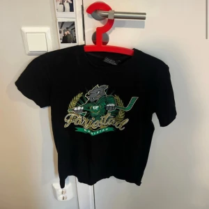 Färjestad t-shirt från South West - Svart t-shirt från South West med Färjestad Karlstad-tryck framtill. Snyggt motiv med varg i grön tröja och hockeyklubba, samt gulddetaljer och text. Klassisk passform och rund hals. Perfekt för dig som älskar hockey och vill visa ditt lagstöd.