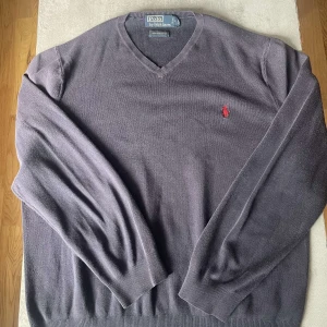 Mörkgrå v-ringad tröja Polo Ralph Lauren - Mörkgrå v-ringad tröja från Polo Ralph Lauren i 100% pima bomull. Klassisk design med röd broderad logga på bröstet och ribbade muddar. Perfekt för dig som gillar stilrena och tidlösa plagg med premiumkänsla.