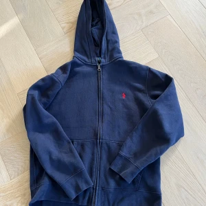 Marinblå hoodie från Polo Ralph Lauren - Snygg marinblå hoodie från Polo Ralph Lauren med dragkedja och huva. Klassisk röd logga broderad på bröstet och stora fickor framtill. Perfekt för dig som gillar en avslappnad och stilren look.