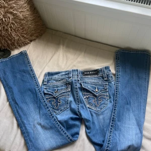 Lågmidjade Rock Revival jeans  - Säljer ett par blå jeans från Rock Revival med vita kontrastsömmar, broderade bakfickor och coola nitar och studs. Lågmidjade bootcut jeans med snygga detaljer och logga på bakfickan. Perfekt för dig som gillar jeans med coola detaljer! Passar någon som har storleken S-M!
