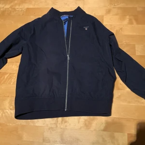 Mörkblå jacka från GANT - Snygg mörkblå vindjacka från GANT med klassisk design och hel dragkedja framtill. Jackan är aldrig använd och är i mycket fint skick! Hör av vid frågor! Pris kan diskuteras