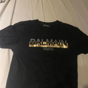 Svart Balmain t-shirt med guldtryck - Svart t-shirt från Balmain med klassisk rund hals och korta ärmar. Framsidan har ett stort Balmain Paris-tryck med en glansig guldremsa som ger en lyxig vibe. Materialet känns mjukt och skönt, perfekt för dig som gillar designerplagg med statement. Bra skick, pris kan diskuteras!
