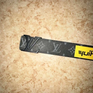   svart bälte med tryck - Svart bälte från med ikoniskt -spänne i matt svart metall. Bältet har ett mönstrat tryck med -loggor och färgglada grafiska detaljer, samt en gul patch med text. Insidan är beige med vit söm. Perfekt för dig som vill sticka ut med designeraccessoar.