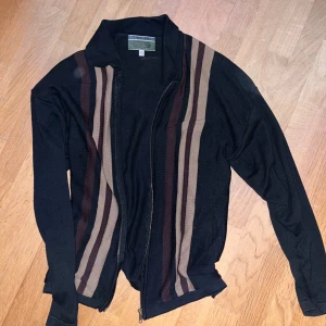 Svart cardigan - Svart kofta från Jackson med dragkedja framtill och coola vertikala ränder i brunt och beige på båda sidor. Långärmad och tillverkad i mjukt stickat material som känns skönt mot huden. Perfekt för dig som gillar retrovibbar och vill sticka ut lite extra.