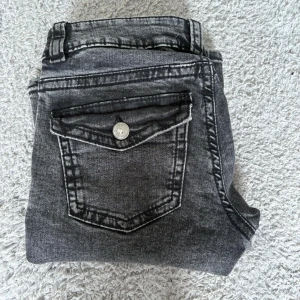 Grå bootcut jeans med fickdetalj - Snygga grå jeans med bootcut-modell och cool ficka med lock och knapp bak. Jeansen har en tvättad look och klassisk femficksdesign. Perfekta för dig som gillar en avslappnad men trendig stil.