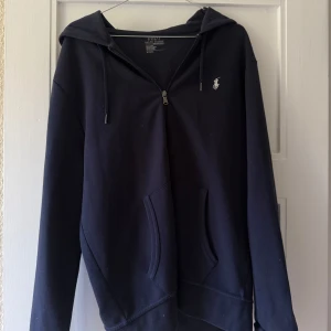 Svart Zip hoodie från Polo Ralph Lauren - Mörkblå hoodie från Polo Ralph Lauren med klassisk logga på bröstet. Tröjan har huva med snörning, hel dragkedja och stora fickor framtill. Tillverkad i mjuk bomull.