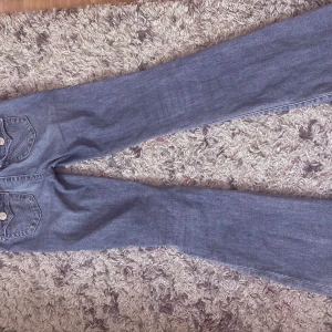 True Religion Becca Bootcut Jeans - Snygga ljusblå bootcut jeans från True Religion, modell Becca Mid Rise. Jeansen har klassiska bakfickor med dekorativa sömmar och silverfärgade knappar. Tillverkade i mjukt denim med en skön passform och medelhög midja.