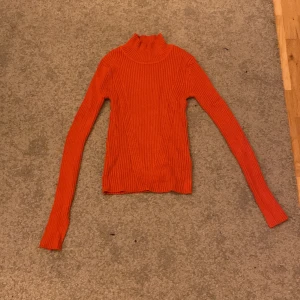 Ribbad orange polotröja - Säljer en ribbad orange/röd polotröja med lång ärm och tight passform. Tröjan är stickad och har en hög krage som ger en snygg siluett. Perfekt att styla med jeans eller kjol för en färgstark look. Har ett håll på ena insida arm, men går enkelt att laga. 